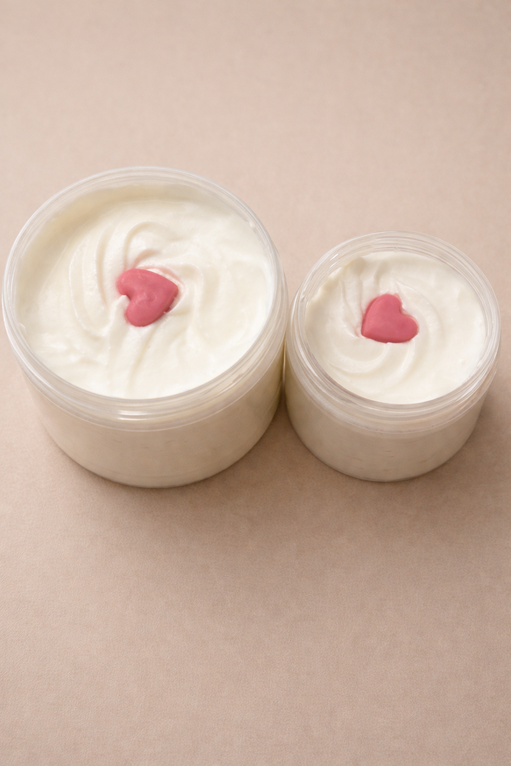 Two containers of white cream body butter with pink heart shapes on a beige background showing how the body butters look when packaged 